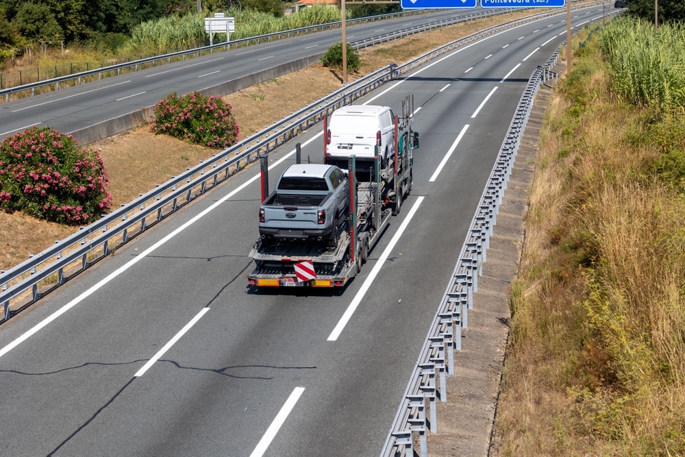Camion porte-voiture : transport de véhicules sur autoroute Camion porte-voiture transportant un Ford Ranger et un fourgon blanc sur une autoroute avec des buissons fleuris.